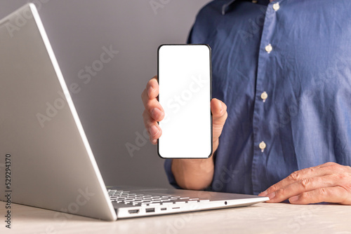 Fototapeta Naklejka Na Ścianę i Meble -  Man showing phone mockup. Male sitting at table with laptop and presenting mobile app at smartphone with white screen. Application advertisement