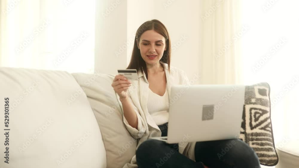 Young attractive woman paying with credit card on laptop laying on the sofa in light living room. Happy customer. Young attractive woman paying with credit card on laptop laying on the sofa in light living room. Happy customer.