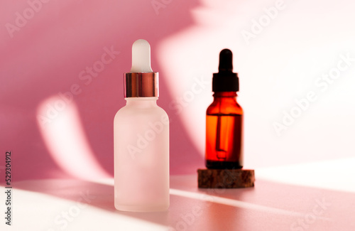 Banner of two transparent amber and frozen glass dropper bottles on pink background in the sunlight. Wooden podium. Pipette with fluid hyaluronic acid, serum, retinol. Cosmetics, healthcare, beauty 