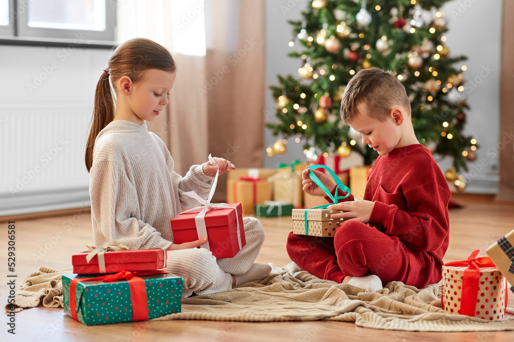 christmas, winter holidays and childhood concept - happy girl and boy in pajamas opening gifts sitting on floor in front of each other at home christmas, winter holidays and childhood concept - happy girl and boy in pajamas opening gifts sitting on floor in front of each other at home