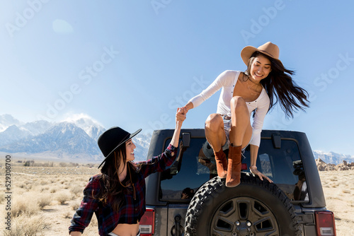 Smiling young woman helping friend to jump from off-road vehicle's spare tire at desert during sunny day