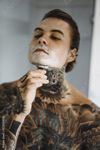 Shirtless tattooed man shaving in bathroom photo