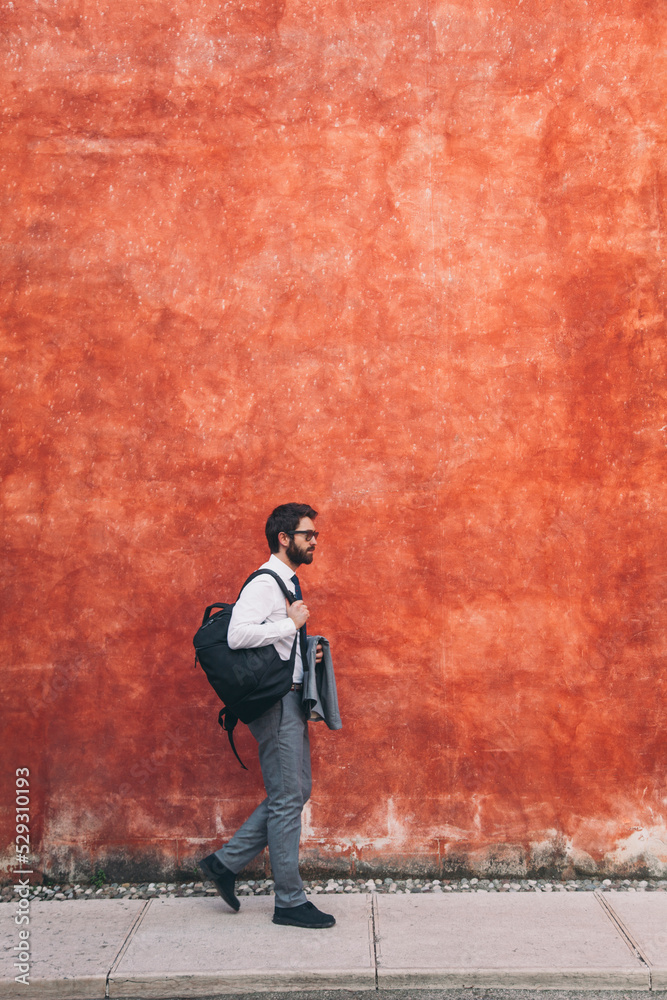 Side view of businessman with backpack walking on sidewalk against wall in city Side view of businessman with backpack walking on sidewalk against wall in city