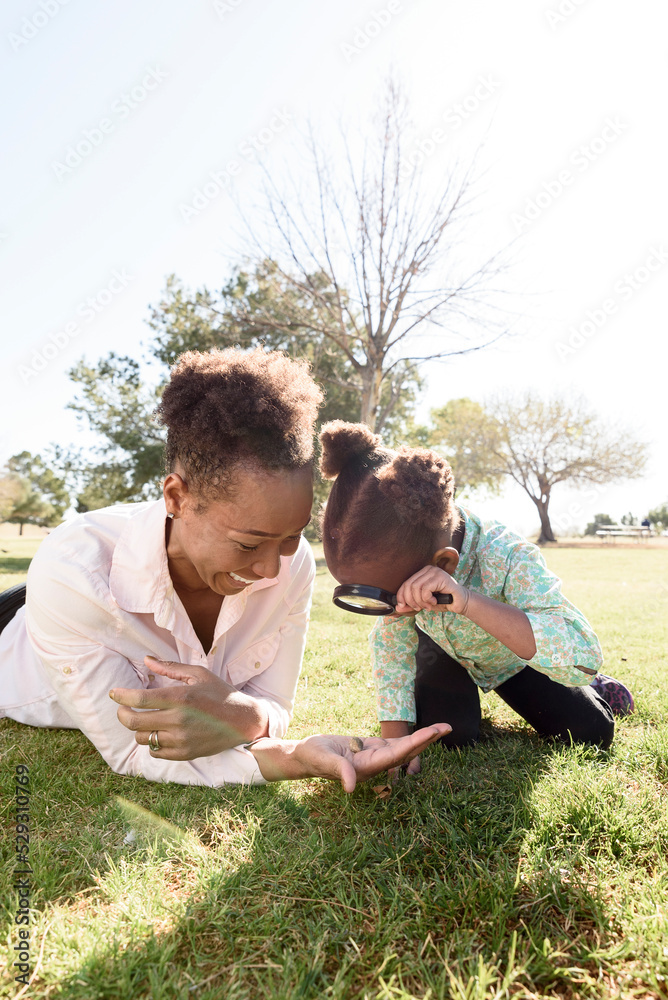 Daughter looking at pebble on mother's hand with magnifying glass in park during sunny day Daughter looking at pebble on mother's hand with magnifying glass in park during sunny day