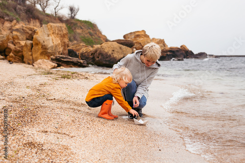 Happy family, friends forever concept. Smiling mother and little son playing with paper boat together near sea in autumn time
