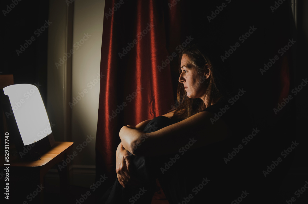 Profile of woman sitting looking at light therapy lamp in a dark room Profile of woman sitting looking at light therapy lamp in a dark room