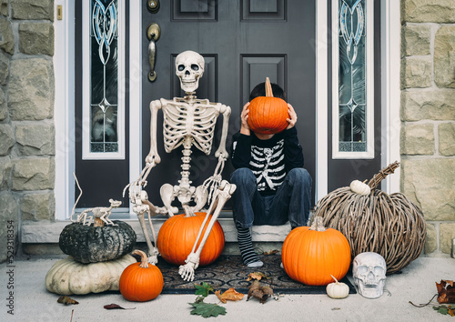 Full length of boy covering face with pumpkin while sitting by skeleton during Halloween