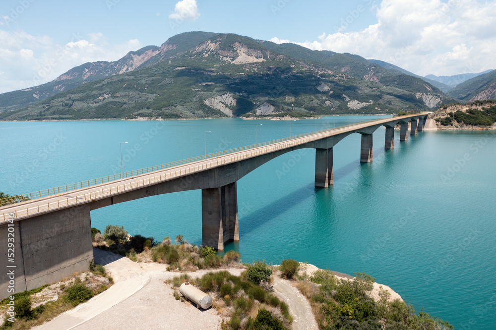 Drone view of bridge over blue lake with mountains at background. Central Greece