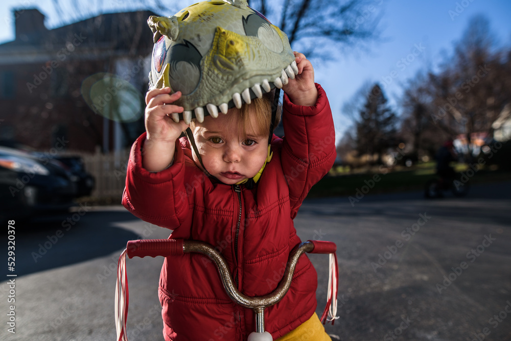 young boy wearing jacket outside on tricycle pulling helmet off head