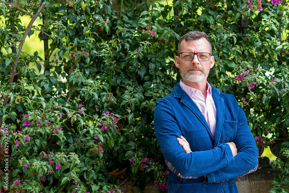 Front view of confident bearded man standing against flowers wall Front view of confident bearded man standing against flowers wall