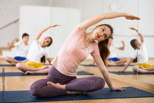 Young woman with her preteen children doing stretching exercises in fitness center, sitting in lotus position and leaning to side with arm outstretched