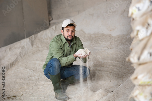 Portrait of focused bearded man farmer crouched down near big pile of maize flour cattle feed, checking quality of forage