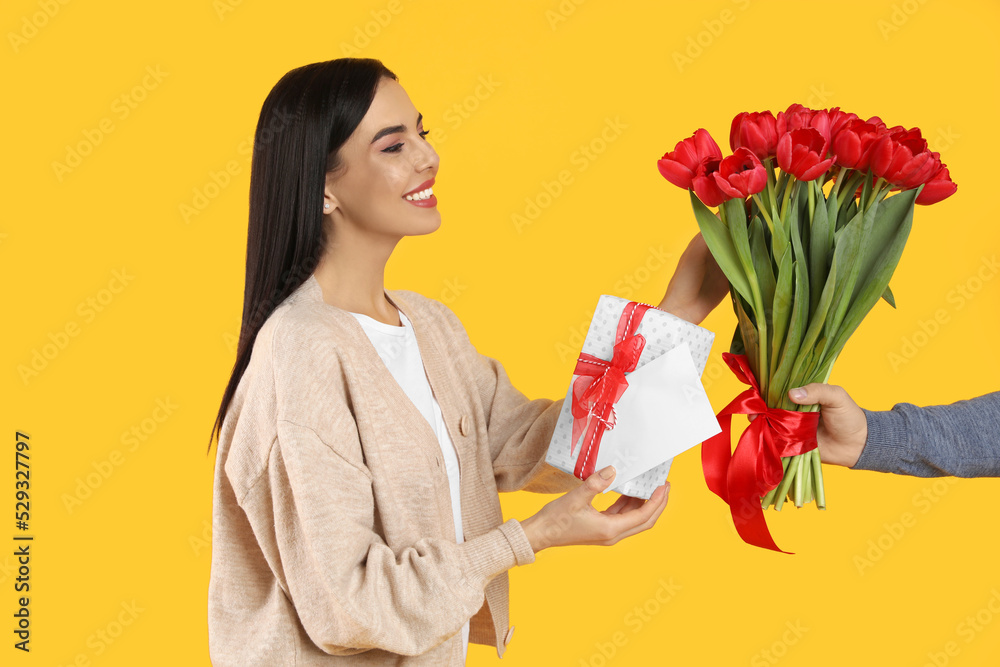 Happy woman receiving red tulip bouquet, greeting card and gift box from man on yellow background. 8th of March celebration Happy woman receiving red tulip bouquet, greeting card and gift box from man on yellow background. 8th of March celebration