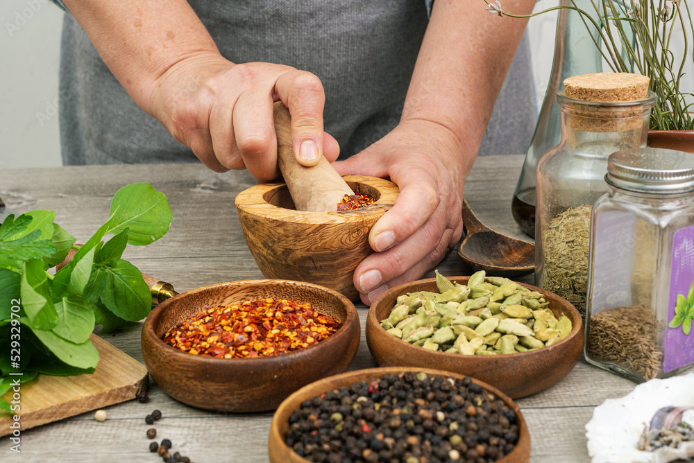 Woman's hands grind red pepper in a wooden mortar. Kitchen gray table with various spices. Woman's hands grind red pepper in a wooden mortar. Kitchen gray table with various spices.