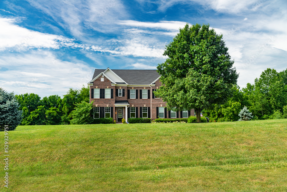 Large traditional red brick colonial house with fluffy wood. Green lawn and blue sky.