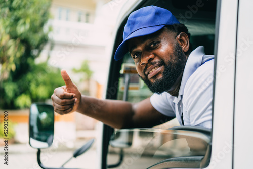 Portrait of courier black man in the truck thumb up and smiling to camera while sitting in driver seat, Optimistic man worker with delivery occupation. photo