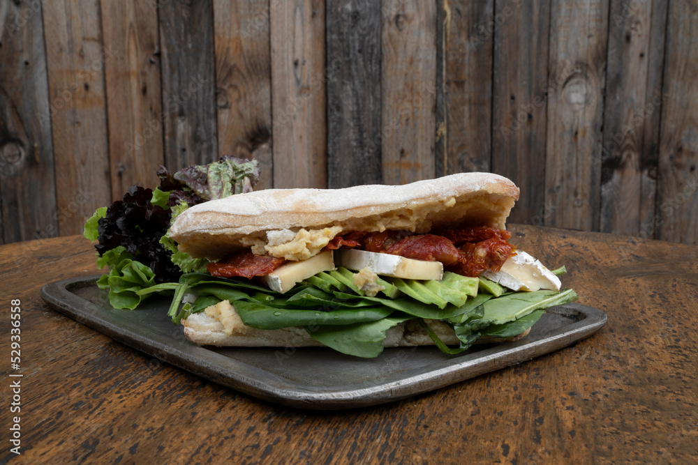 Obraz premium Gourmet sandwich. Closeup view of vegetarian sandwich with ciabatta bread, chickpea hummus, avocado, sun dried tomatoes, brie cheese and spinach, in a metal dish with a wooden background. 