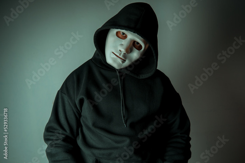 A portrait of an anonymous hacker wearing a mask and a black hoodie sitting with his head tilted and terrifying. Hacking and malware concept.