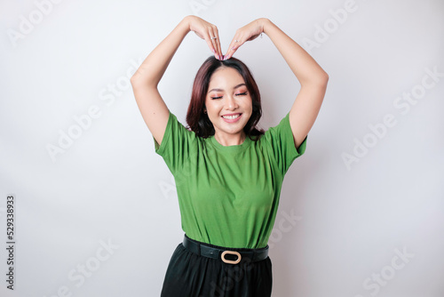 An attractive young Asian woman wearing a green t-shirt feels happy and a romantic shapes heart gesture expresses tender feelings © Reezky
