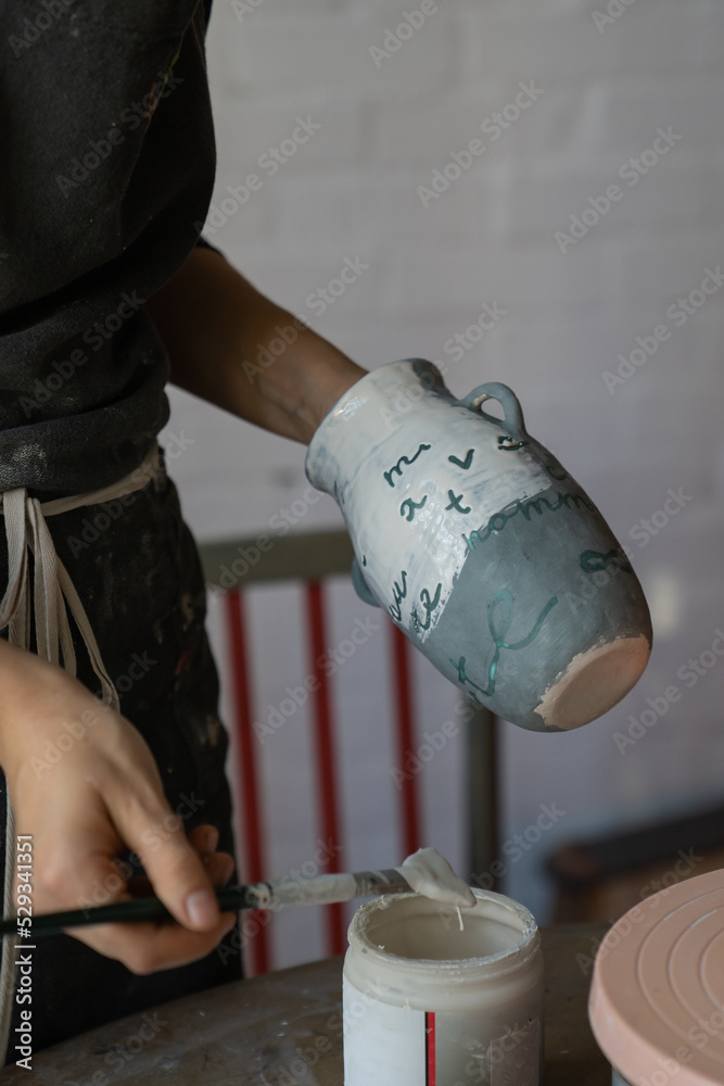 Woman in apron works with pottery standing near round wooden table with equipment. Professional female artisan creates handmade ceramic tableware painting white vase with brush in workshop closeup