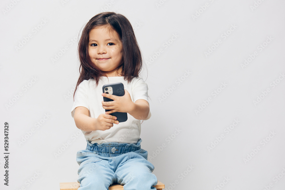 a cute little preschool girl is standing on a white background in a white T-shirt, sitting on a chair, holding a phone in her hands and smiling. The theme of children's happiness a cute little preschool girl is standing on a white background in a white T-shirt, sitting on a chair, holding a phone in her hands and smiling. The theme of children's happiness