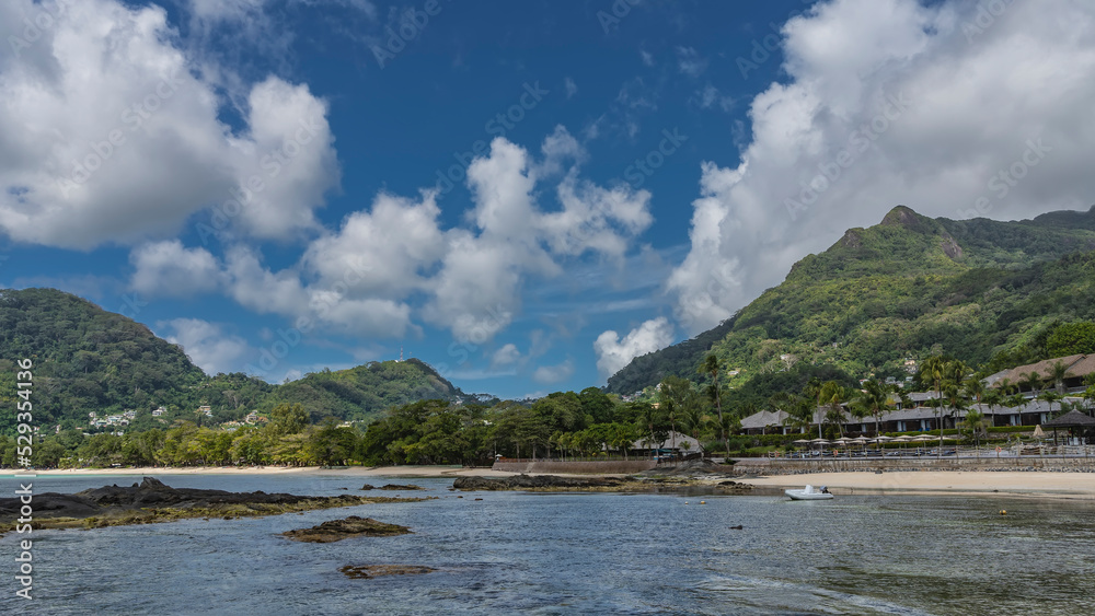 On the ocean shore, in the recreation area, canopies, sun umbrellas are visible. Green mountains against a background of blue sky and clouds. A white boat is moored at the shore. Seychelles. Mahe