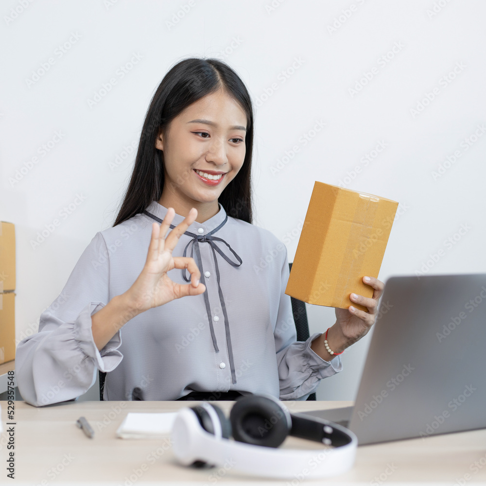 Young Asian woman chatting with a customer on a laptop and displays the product before delivery to confirm the order, Selling products online or doing freelance work at home concept. Young Asian woman chatting with a customer on a laptop and displays the product before delivery to confirm the order, Selling products online or doing freelance work at home concept.