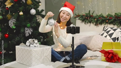 Young smiling asian woman with santa hat and xmas bauble in hand speaking on phone via video link at home, holiday time, festive decoration of bedroom interior by Christmas tree and lights