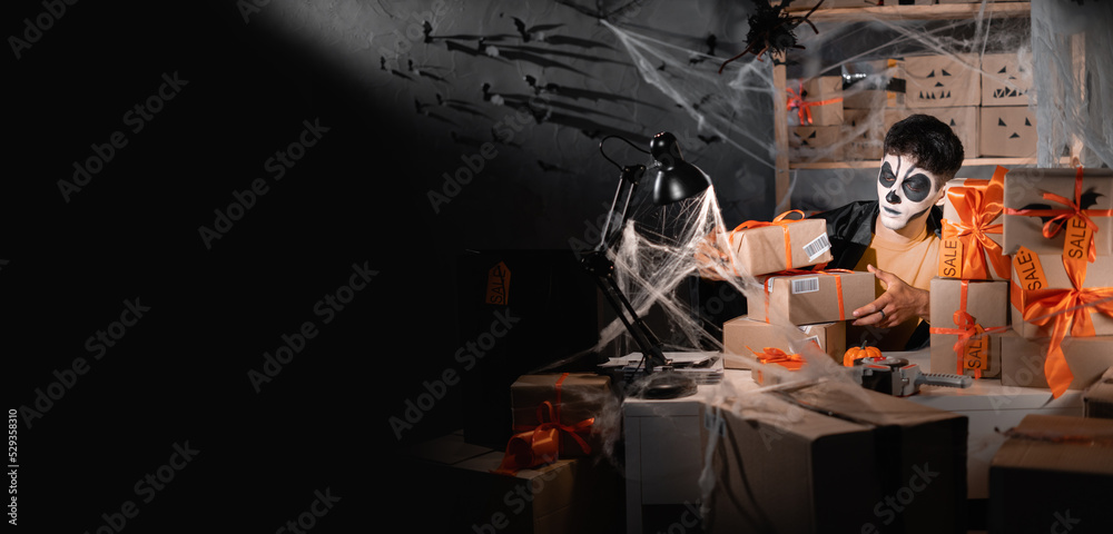 start up small business owner man wear white clay skull, professional make up in home office spider and cobweb preparing parcel to send. Banner.