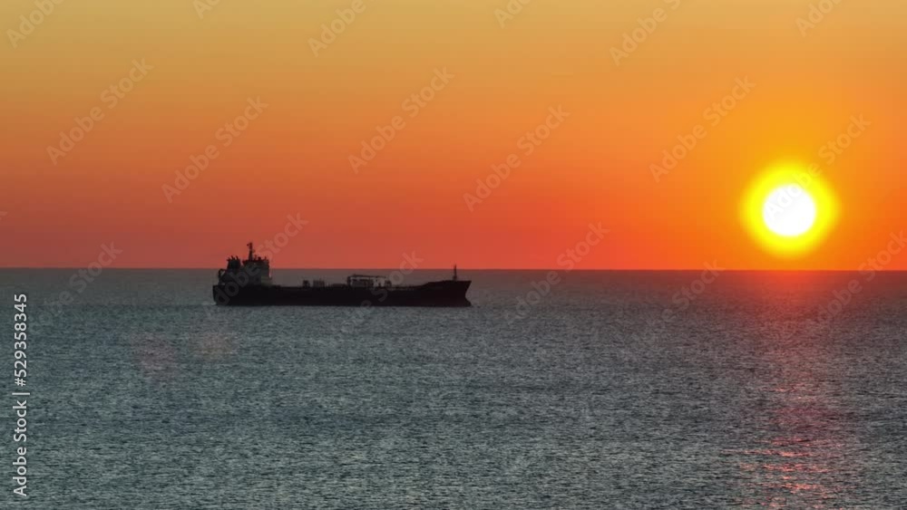 Large merchant ship sailing through sea aerial view. Cargo is sent to city's seaport receive unload cargo at sunrise in morning in winter. Import, export. Bright disc of pink dawn sun.