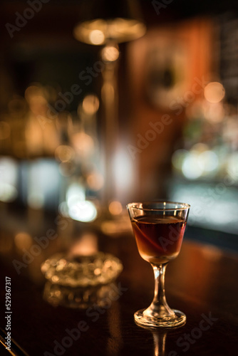 A glass with cold Manhattan cocktail garnished with orange zest peel on a wooden bar counter. Bokeh lights, selective focus, small depth of field