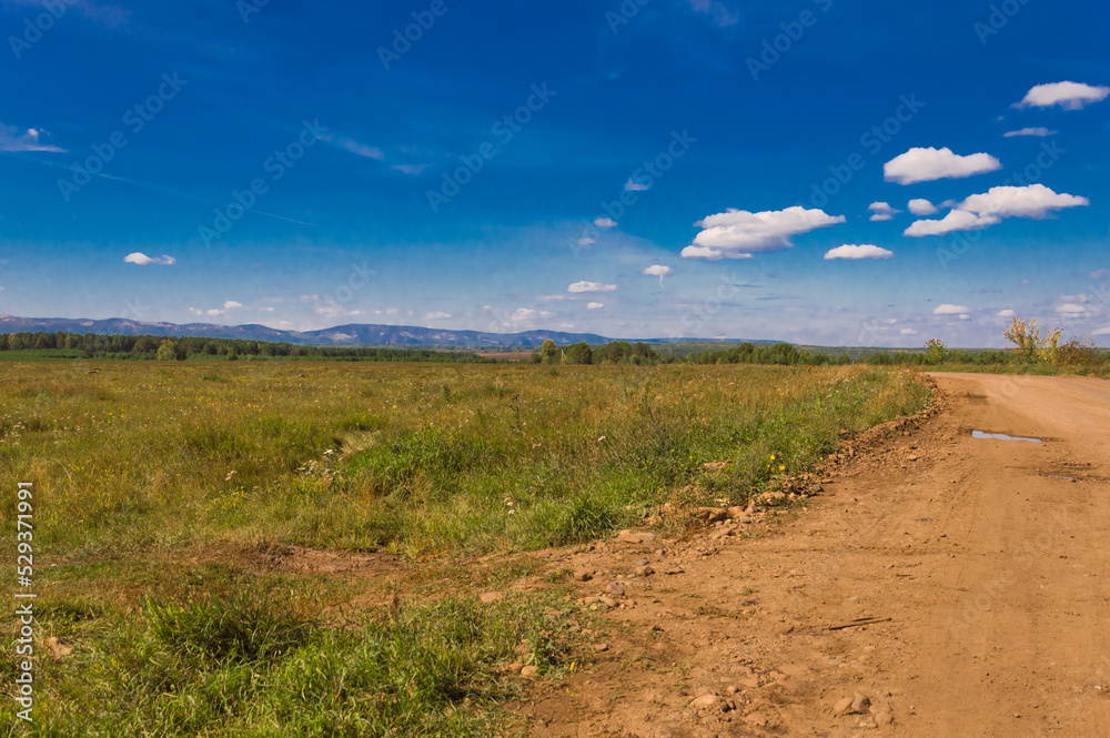 Landscape. Country road with red soil. Side meadow with grass. On the horizon are the blue mountains of the Sayan Mountains. Eastern Siberia. Beautiful blue sky with clouds.