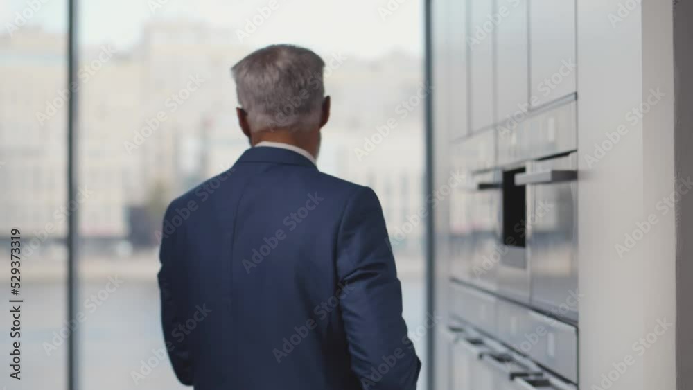 Mature man in suit take bottle of juice from fridge and drink standing near large window. Realtime Mature man in suit take bottle of juice from fridge and drink standing near large window. Realtime