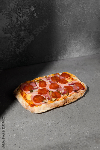 Traditional italian meat pizza with pepperoni and bacon on dark background. Meat pinsa on gray stone table with hard shadow. Roman pizza with pepperoni and bacon on concrete background. Pinsa menu.