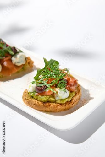 Aesthetic composition with salmon bruschetta on white background with shadows from flowers. Italian bruschetta with salmon  avocado  cheese and olives on fine dining in summer. Elegant menu concept.