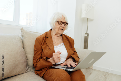 a serious, experienced, elderly woman is sitting working from home on the couch with a laptop on her lap and menacingly looks at the monitor with her hand in a fist