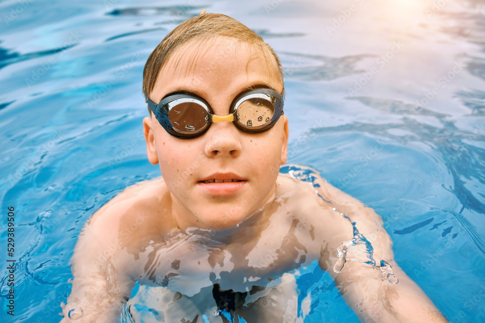 Naklejka premium Smiling boy portrait in swimming goggles, Child swim in the pool, sunbathes, swimming in hot summer day. Relax, Travel, Holidays, Freedom concept. 4K slow motion
