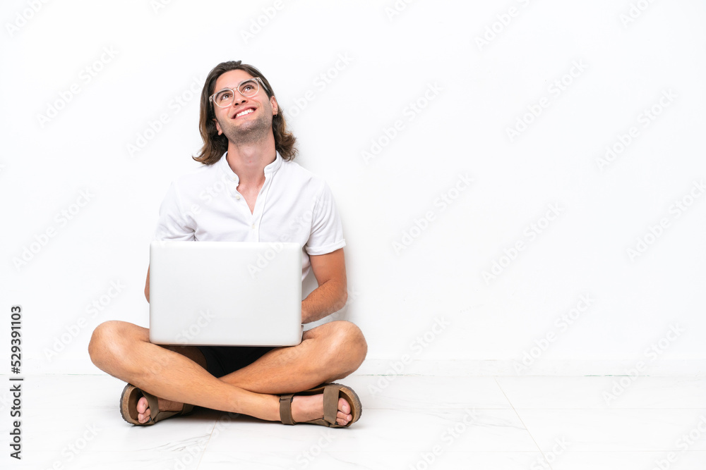 Young handsome man with a laptop sitting on the floor isolated on white background looking up while smiling Young handsome man with a laptop sitting on the floor isolated on white background looking up while smiling