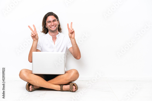 Young handsome man with a laptop sitting on the floor isolated on white background showing victory sign with both hands © luismolinero