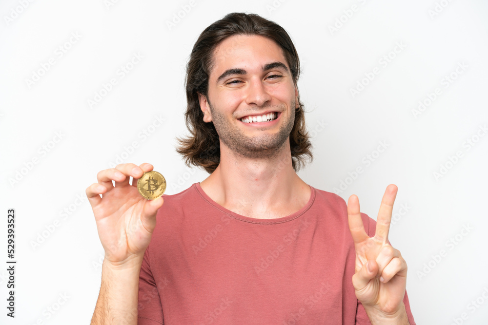 Young handsome man holding a Bitcoin isolated on white background smiling and showing victory sign Young handsome man holding a Bitcoin isolated on white background smiling and showing victory sign