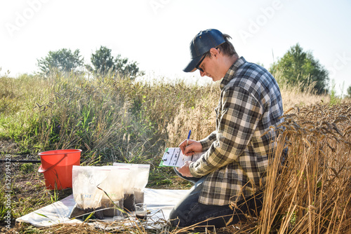 Male agronomist preparing soil samples in sample bags for laboratory analysis, writing in information sheet outdoors at sunrise. Man farmer taking notes in form, working at field. Soil certification