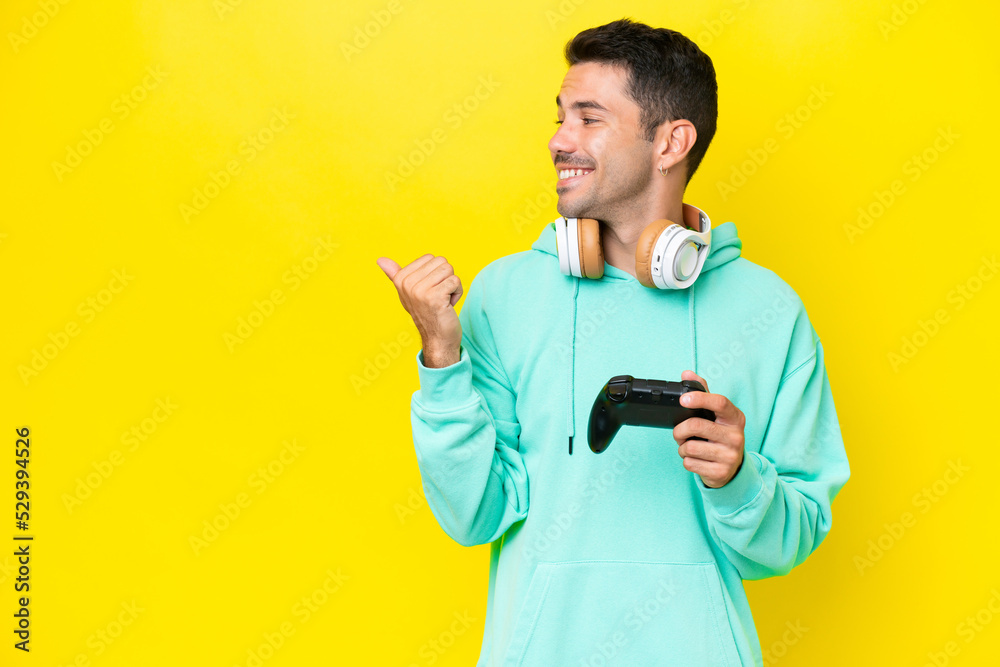Young handsome man playing with a video game controller over isolated wall pointing to the side to present a product Young handsome man playing with a video game controller over isolated wall pointing to the side to present a product