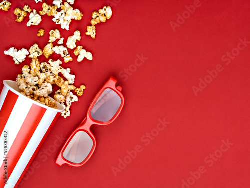 Popcorn spilled from a white and red striped bucket near 3D glasses on a red background. Top view, above, overhead. Space for text. Cinema, movies, movie night and entertainment concept.