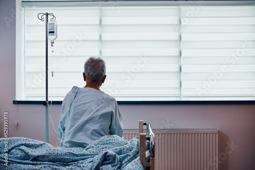 Fotografie, Tablou Rear view of senior  male patient with IV drip sitting on bed while recovering in hospital ward