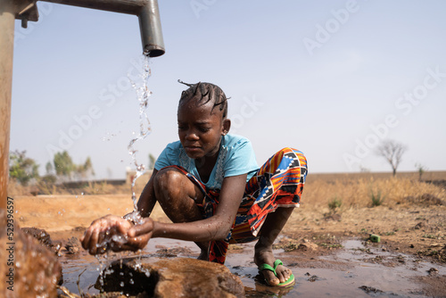 A young African girl casually crouches in front of a water pump and washes roughly  which is symbolic of the lack of tap water and washing facilities in the developing world