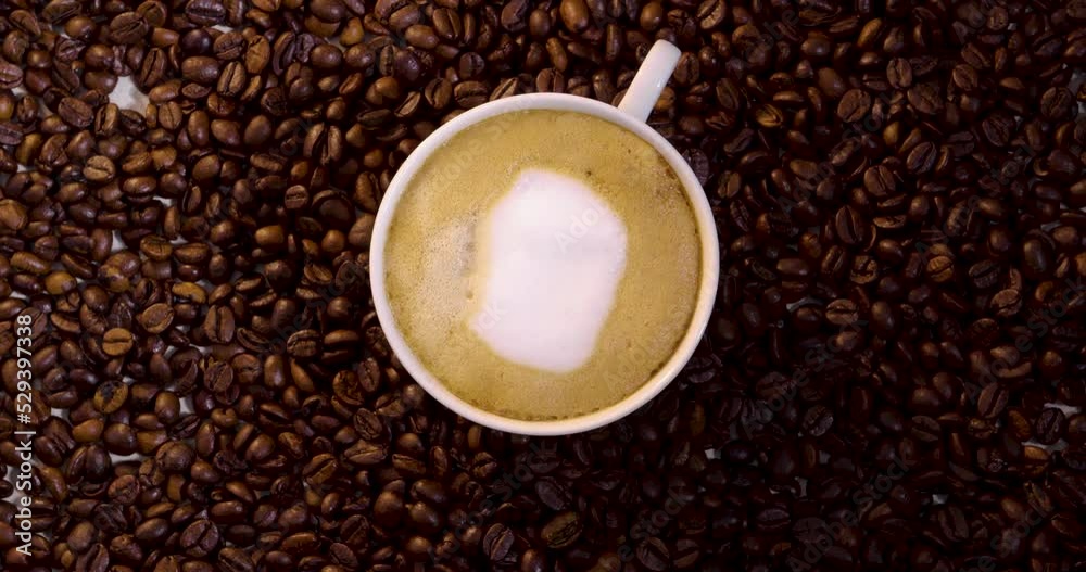 Birdseye view of a spinning cup filled with foamy coffee latte and cream surrounded by a pile of coffee beans Birdseye view of a spinning cup filled with foamy coffee latte and cream surrounded by a pile of coffee beans