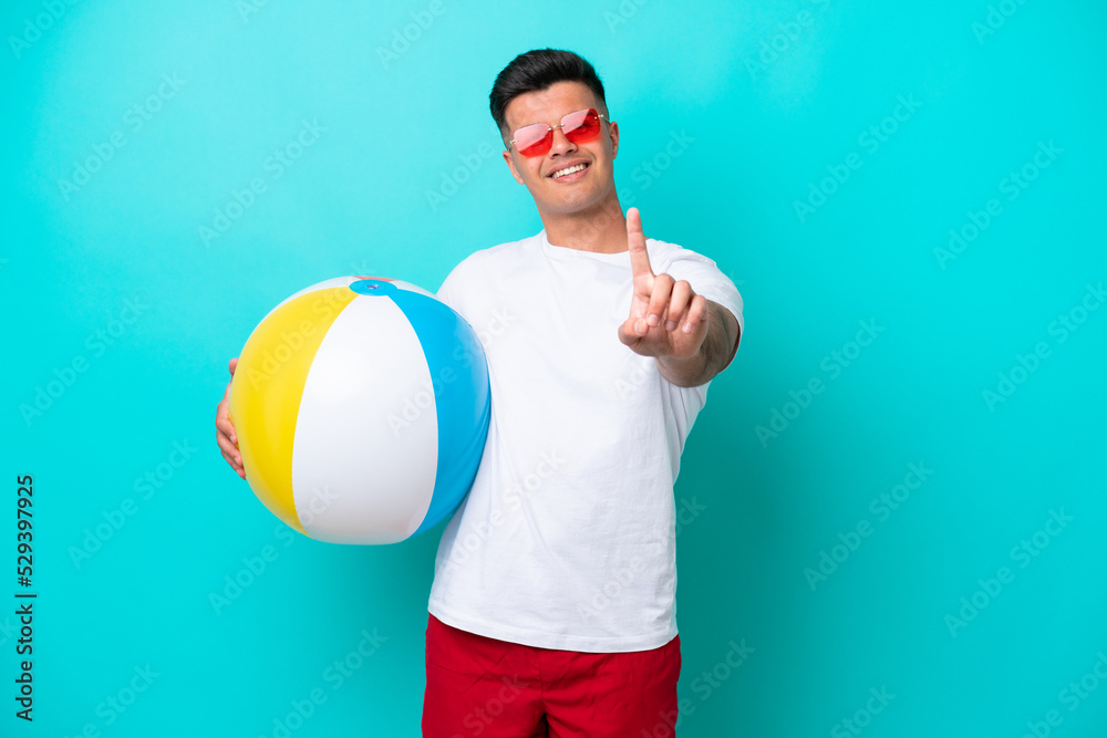 Young caucasian man holding a beach ball isolated on blue background showing and lifting a finger