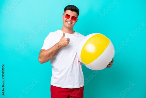 Young caucasian man holding a beach ball isolated on blue background giving a thumbs up gesture © luismolinero