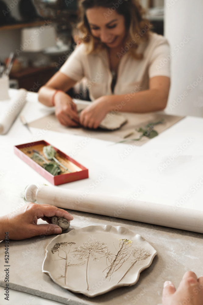 Woman decorating with flowers rolled clay, making ceramic plate in studio with floral pattern. Handmade creative work. Pottery workshop for adults. Woman decorating with flowers rolled clay, making ceramic plate in studio with floral pattern. Handmade creative work. Pottery workshop for adults.