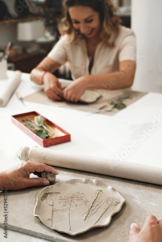 Woman decorating with flowers rolled clay, making ceramic plate in studio with floral pattern. Handmade creative work. Pottery workshop for adults. 